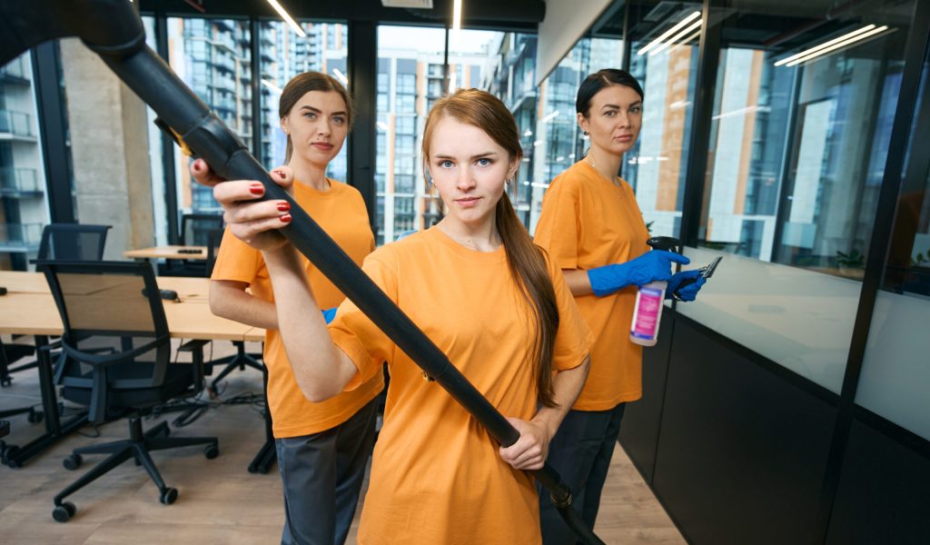 Three cleaning company workers in uniform and protective gloves cleaning and disinfecting office space with vacuum cleaner, rag and spray